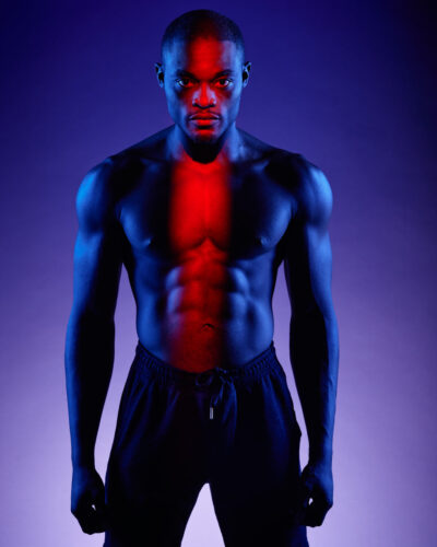 male modeling photography tips - Chido stands shirtless, illuminated with dramatic red and blue lighting that highlights his muscular physique and creates a striking visual contrast in this 3/4 length portrait