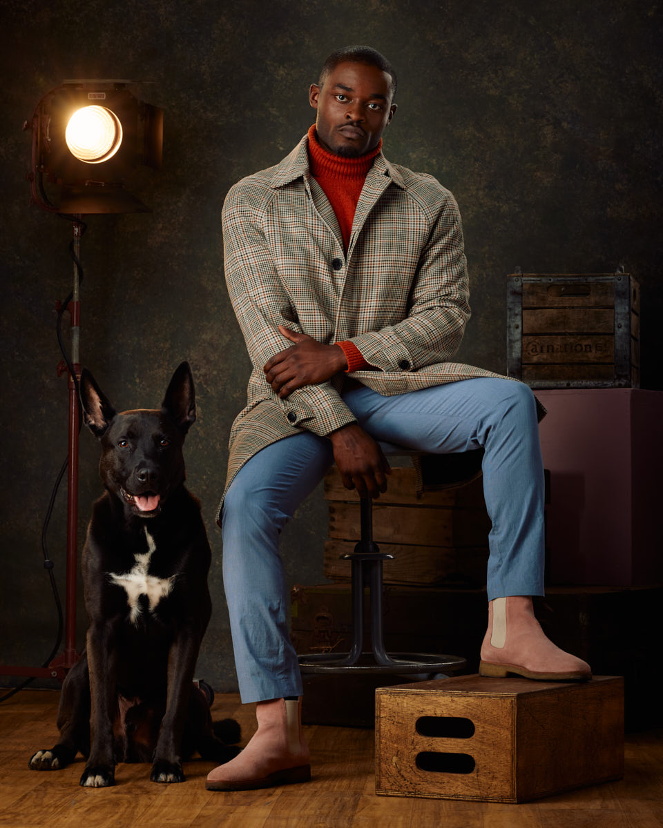 Chido sits on a stool in a studio setting, dressed in a plaid coat, red turtleneck, blue pants, and brown suede shoes, with a black dog sitting next to him and a vintage studio light illuminating the scene.
