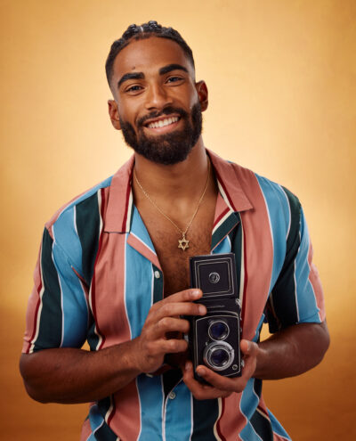 Shomari stands in front of a warm, golden background, holding a vintage camera. He is dressed in a colorful, striped short-sleeved shirt, and wears a Star of David necklace. His broad smile and direct gaze create a welcoming and charismatic portrait, reflecting a blend of modern style and nostalgic charm.