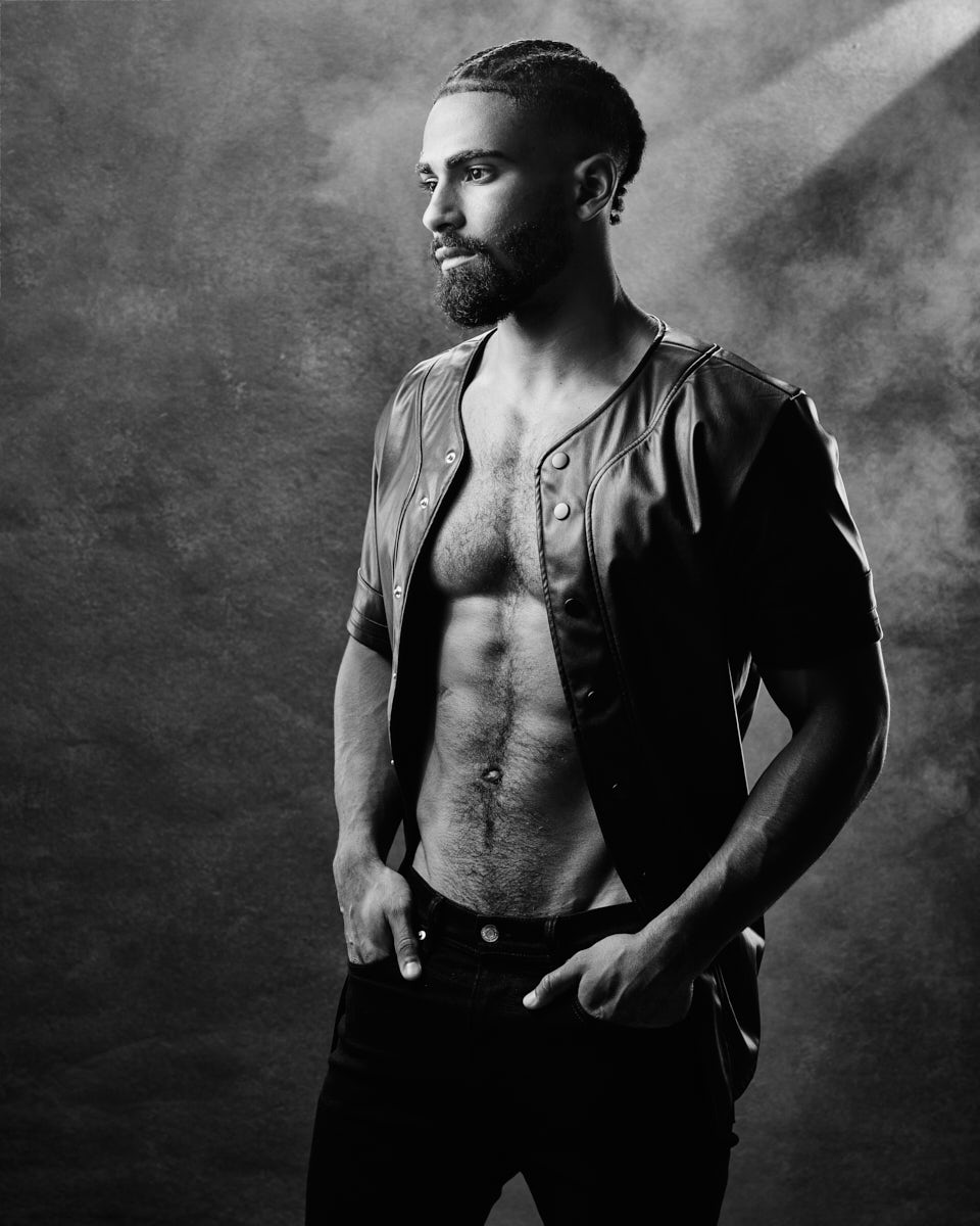 A black and white photo of Shomari, shirtless and wearing an open leather jacket. He stands against a textured background, looking off to the side, with his hands in his pockets, conveying a rugged, contemplative look.