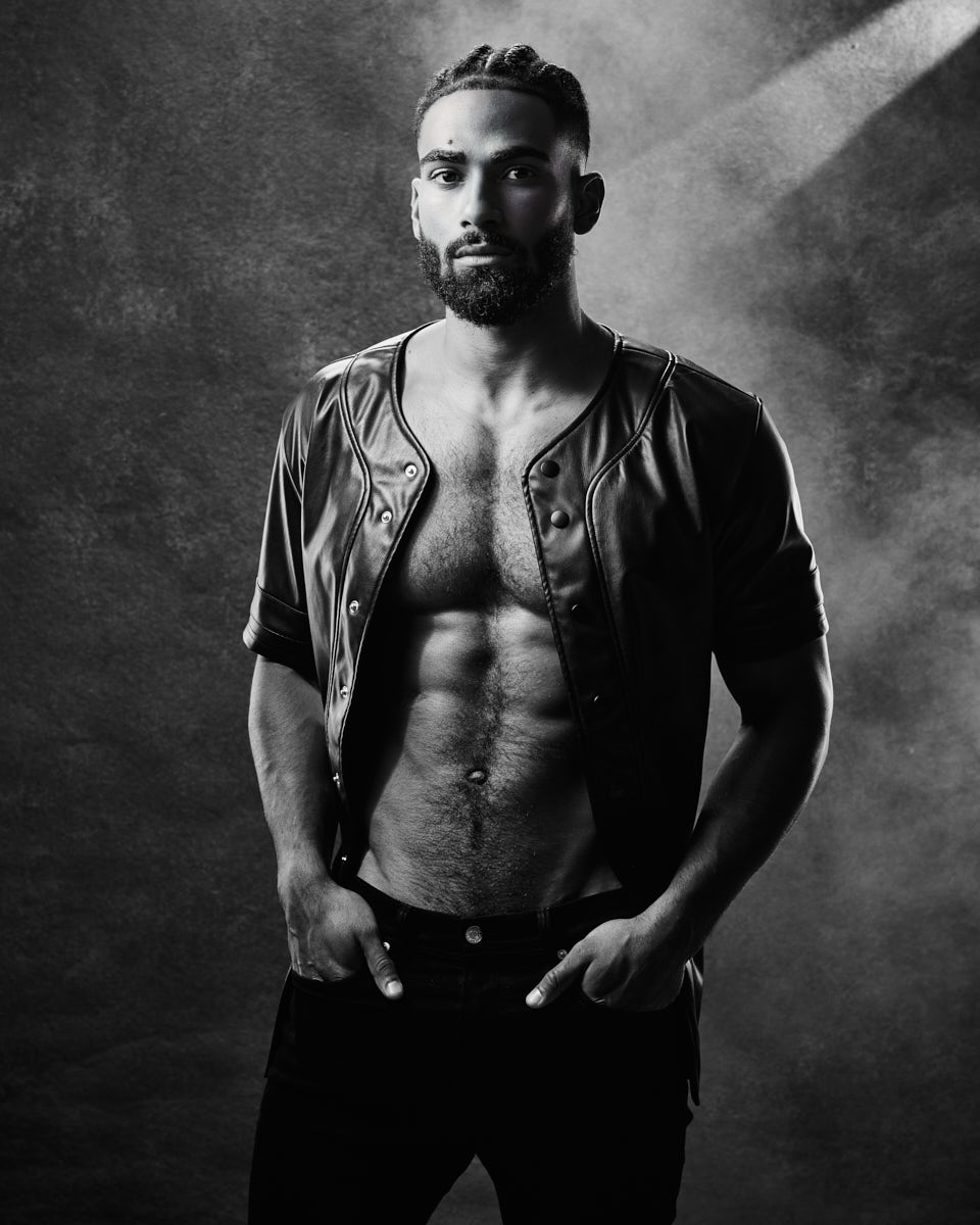 Another black and white portrait of Shomari, shirtless with an open leather jacket. He faces the camera with a serious expression, hands in his pockets, and the lighting emphasizes his muscular build.