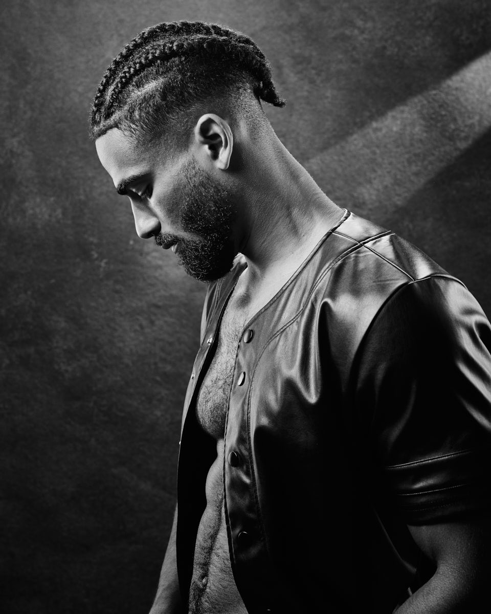 Shomari poses in a dramatic black and white close-up. The focus is on his profile, showcasing his braided hairstyle and strong facial features against a dark background.
