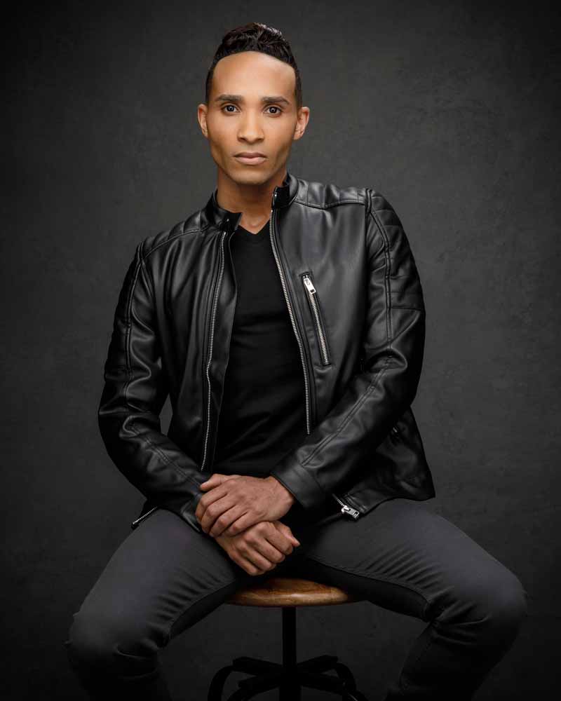 Anthony is sitting on a wooden stool, wearing a black leather jacket and a black shirt, looking directly into the camera with a confident expression.