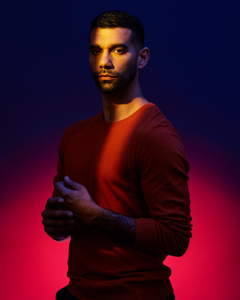 Fernando is posing against a dark blue and pink background, lit by dramatic lighting that casts a strong yellow beam across his face and upper body. He is wearing a red sweater and looking directly at the camera with a serious expression. His hands are held together in front of him, and a tattoo is visible on his arm.