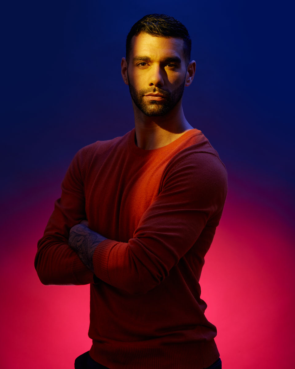 Similar to the first image, Fernando stands with his arms crossed against the same background. The lighting again creates a striking effect with a beam of light illuminating his face. He maintains a confident and intense gaze towards the camera.