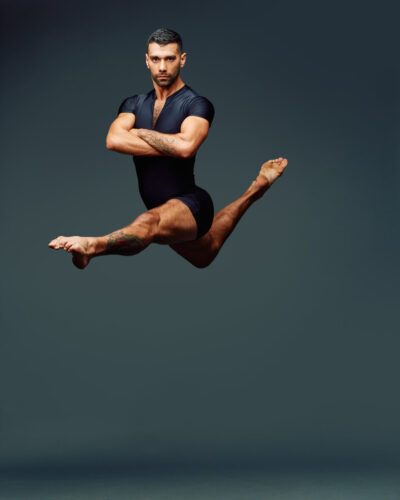 Another action shot of Fernando mid-air, this time with his arms crossed over his chest. His legs are in a split position, and he maintains a focused expression. His tattoo is visible on his leg, and the image captures the height and grace of his jump.