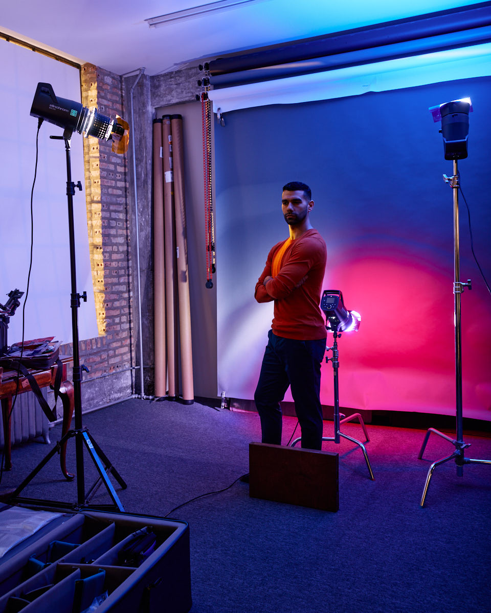 Fernando stands in a studio setup, with lighting equipment and backdrop visible. He is wearing the same red sweater and dark pants as in the earlier images. He stands with his arms crossed, looking directly at the camera with a composed expression. The background lighting creates a colorful and vibrant effect.