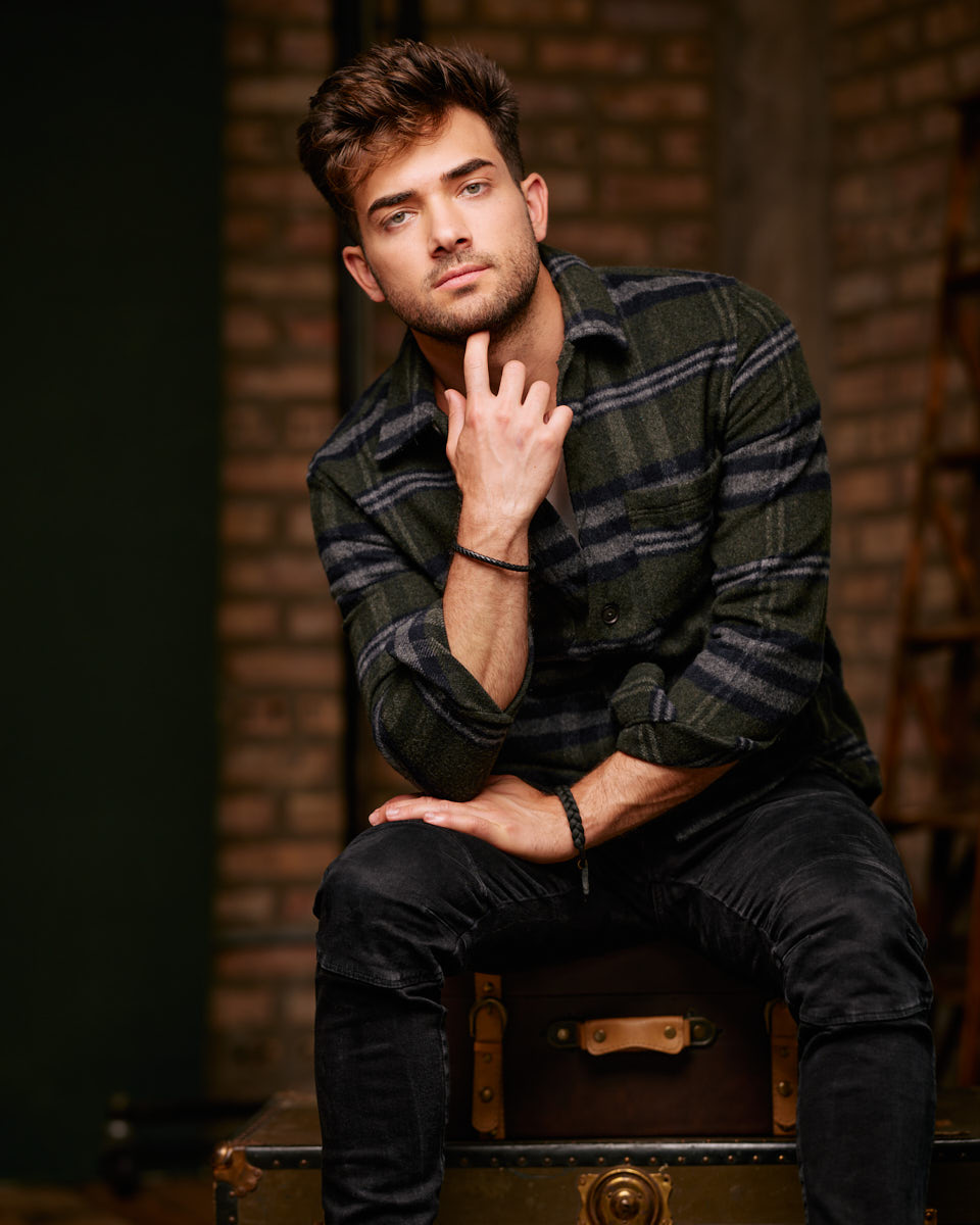 Chicago editorial lookbook photographer -Eric sitting on a vintage trunk, wearing a green and black plaid shirt, with a thoughtful expression, set against a brick wall.