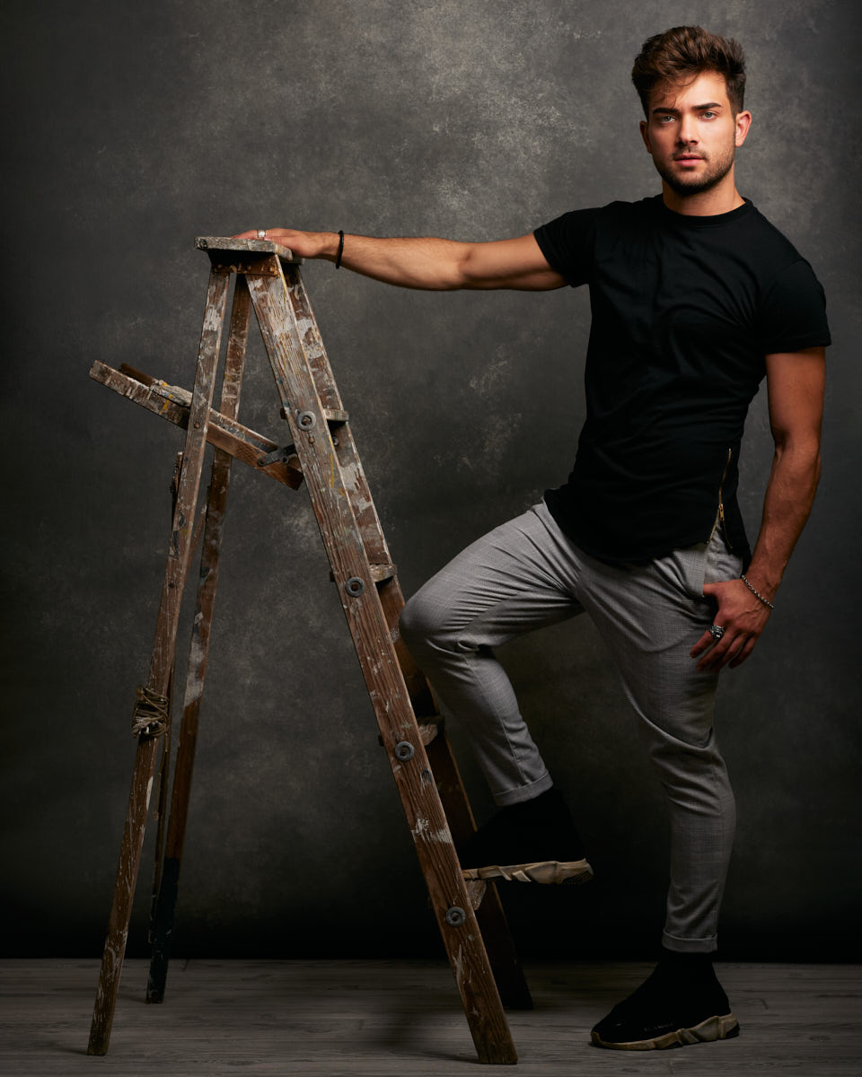 Chicago editorial lookbook photographer -Eric is dressed in a black t-shirt and gray pants, posing with one foot on a wooden ladder, exuding a strong, confident vibe.