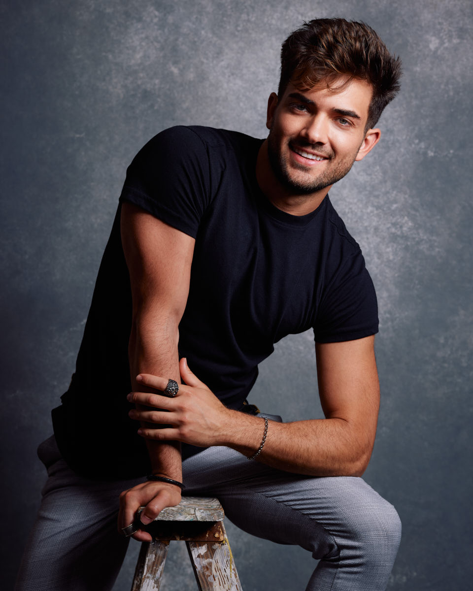 Chicago editorial lookbook photographer -Eric in a black t-shirt and gray pants, smiling and leaning on a wooden stool, showcasing a friendly and approachable demeanor.