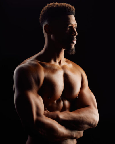 Side profile of Pierre, shirtless, showcasing his muscular upper body against a dark background, emphasizing his fitness and strength.