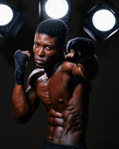 Kyle, a muscular male model in a boxing pose, with fists up and wrapped in hand wraps, under dramatic studio lighting highlighting his defined physique.