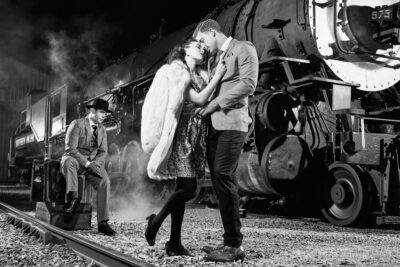 Derrick and Alina in a close embrace near the train, with Jake seated on a suitcase, contributing to a romantic and mysterious atmosphere.