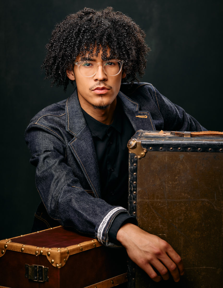 Chicago male fashion model headshots - Kasan leans on a stack of vintage suitcases, wearing a denim jacket and glasses. His serious expression and the setup create a sophisticated look.