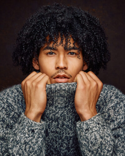 A close-up of Kasan with his hands pulling up his sweater collar, emphasizing his eyes and facial features. The texture of the sweater adds depth to the portrait.