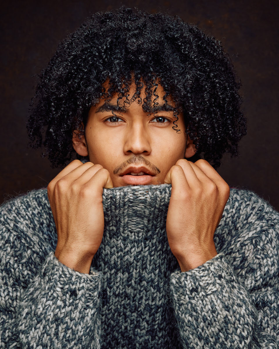 A close-up of Kasan with his hands pulling up his sweater collar, emphasizing his eyes and facial features. The texture of the sweater adds depth to the portrait.
