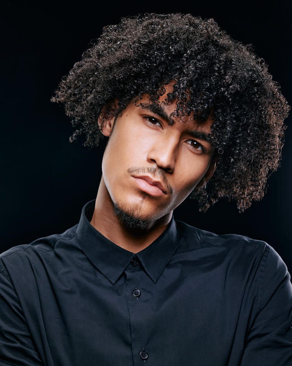 Another headshot of Kasan, tilting his head slightly, wearing a black shirt. The dark background emphasizes his facial features and curly hair.