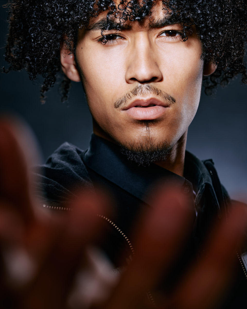 A close-up of Kasan’s face, with his hand reaching towards the camera, creating a sense of depth. His serious expression and focus make the image compelling.