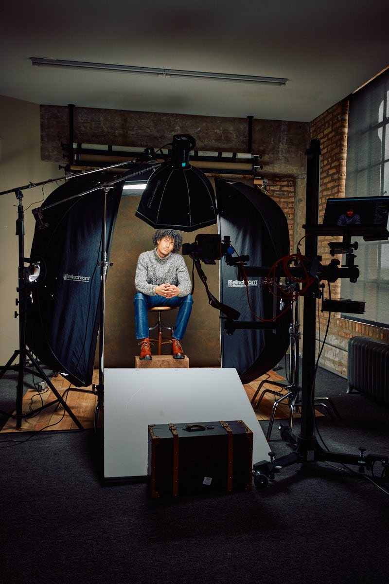 Behind the scene of Kasan sitting on a stool, dressed in a chunky gray knit sweater and jeans. He is surrounded by professional studio lighting equipment and reflectors, indicating a photoshoot setup. His relaxed posture and serious expression add a thoughtful mood to the scene.