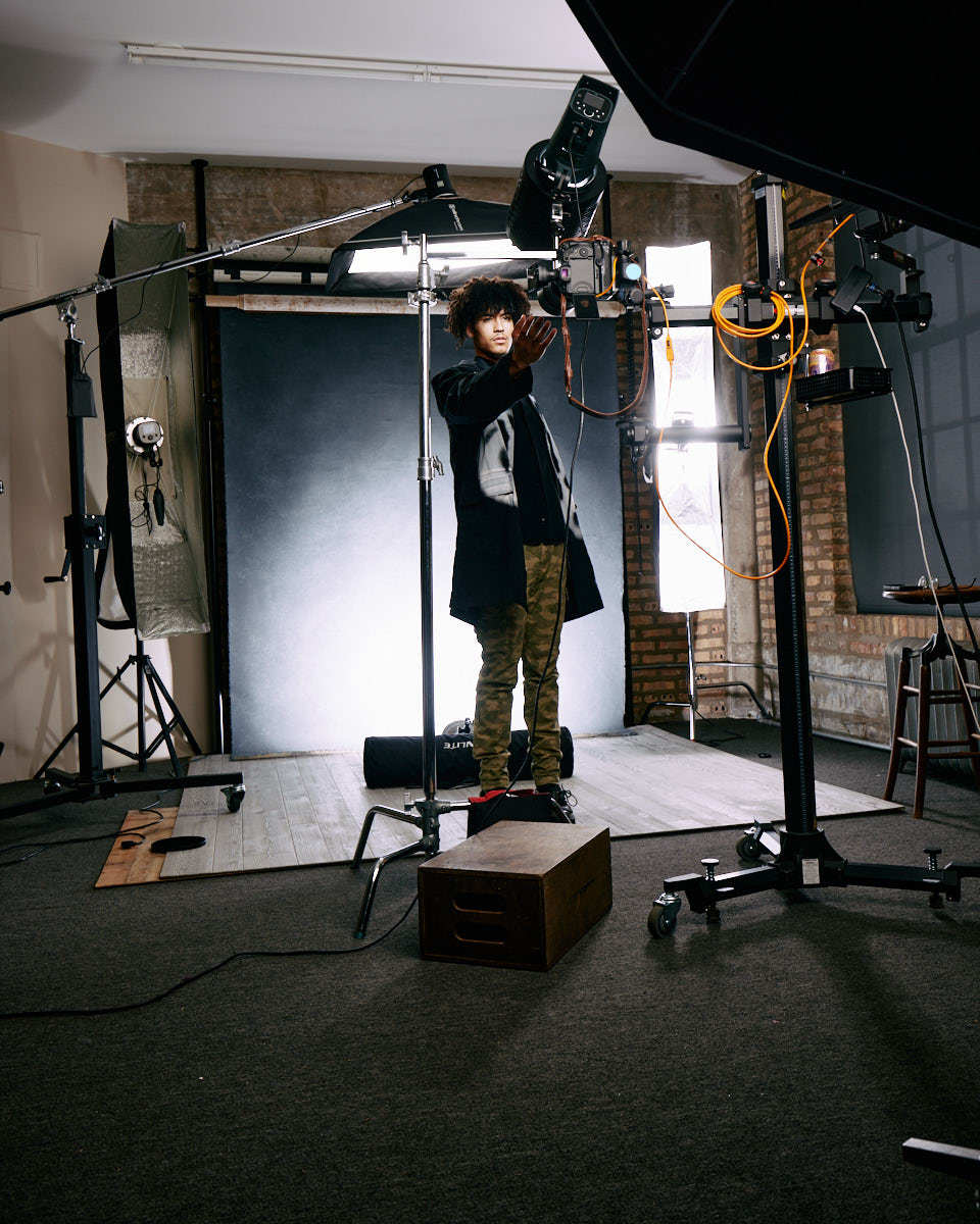 Wearing a dark jacket and camo pants, Kasan stands in a studio setting. He is adjusting the lighting equipment, which emphasizes the behind-the-scenes aspect of the photoshoot. The image captures the technical environment of a professional photography session.