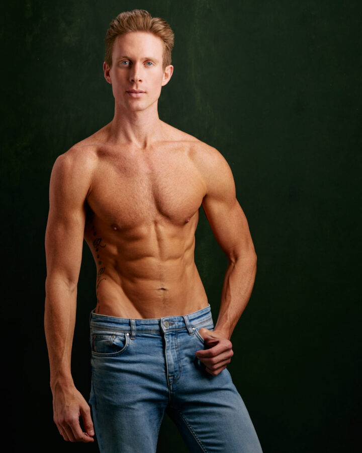 Lucas posed shirtless against a dark green background, showing off his well-defined, muscular physique. He is wearing light blue jeans and looking directly at the camera with a serious expression.