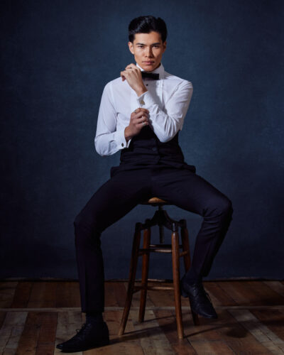 Santiago sits on a stool, adjusting his bow tie with a composed and elegant demeanor, emphasizing his formal and sophisticated side.