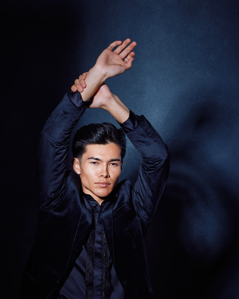 Santiago stands against a dark blue background with his arms gracefully crossed above his head, showcasing his flexibility and elegant posture