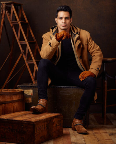 Paulo sitting in a studio setup, wearing a tan coat, black jeans, and brown gloves, looking relaxed yet stylish with a rustic backdrop.