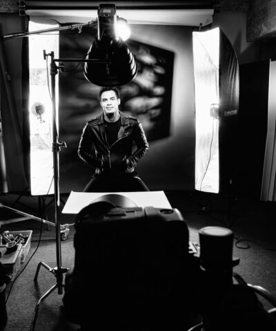 Behind-the-scenes shot of Paulo in a studio, sitting in a leather jacket with a lighting setup visible, highlighting the professional shoot environment.