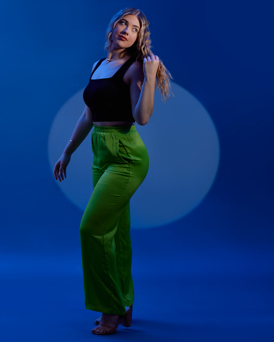 Editorial Model Photography in Chicago- A model wearing a black crop top and bright green high-waisted pants, posing with her right hand touching her hair, standing against a blue background with a circular light effect.