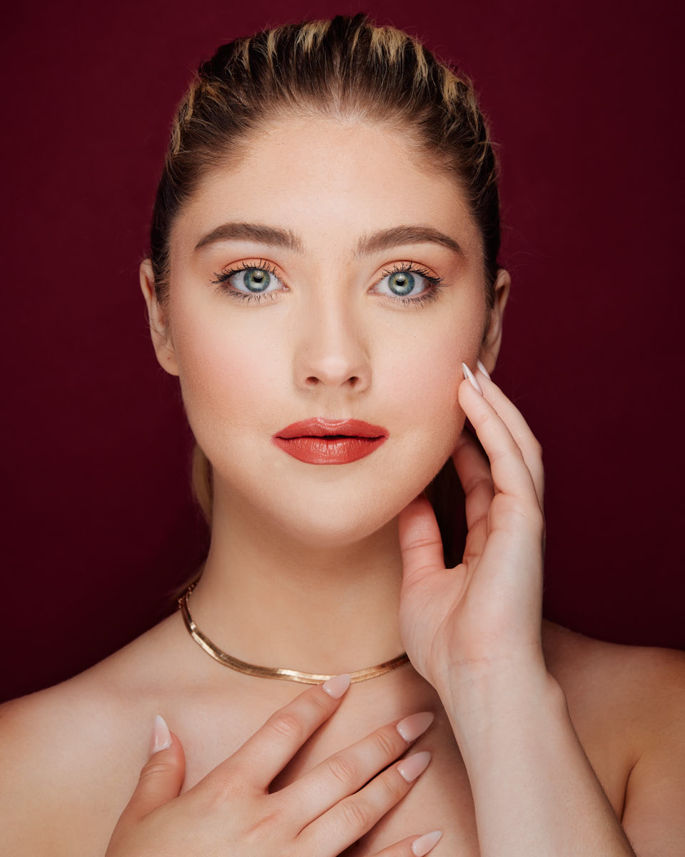 Editorial Model Photography in Chicago-A close-up portrait of the same model with blue eyes, wearing a gold choker and red lipstick, posing against a burgundy background.