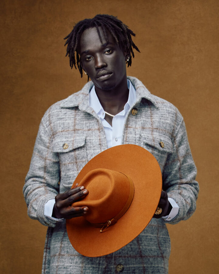 Jada holds an orange hat in front of his chest, wearing a gray plaid overcoat over a white shirt. The background is a warm, textured brown.