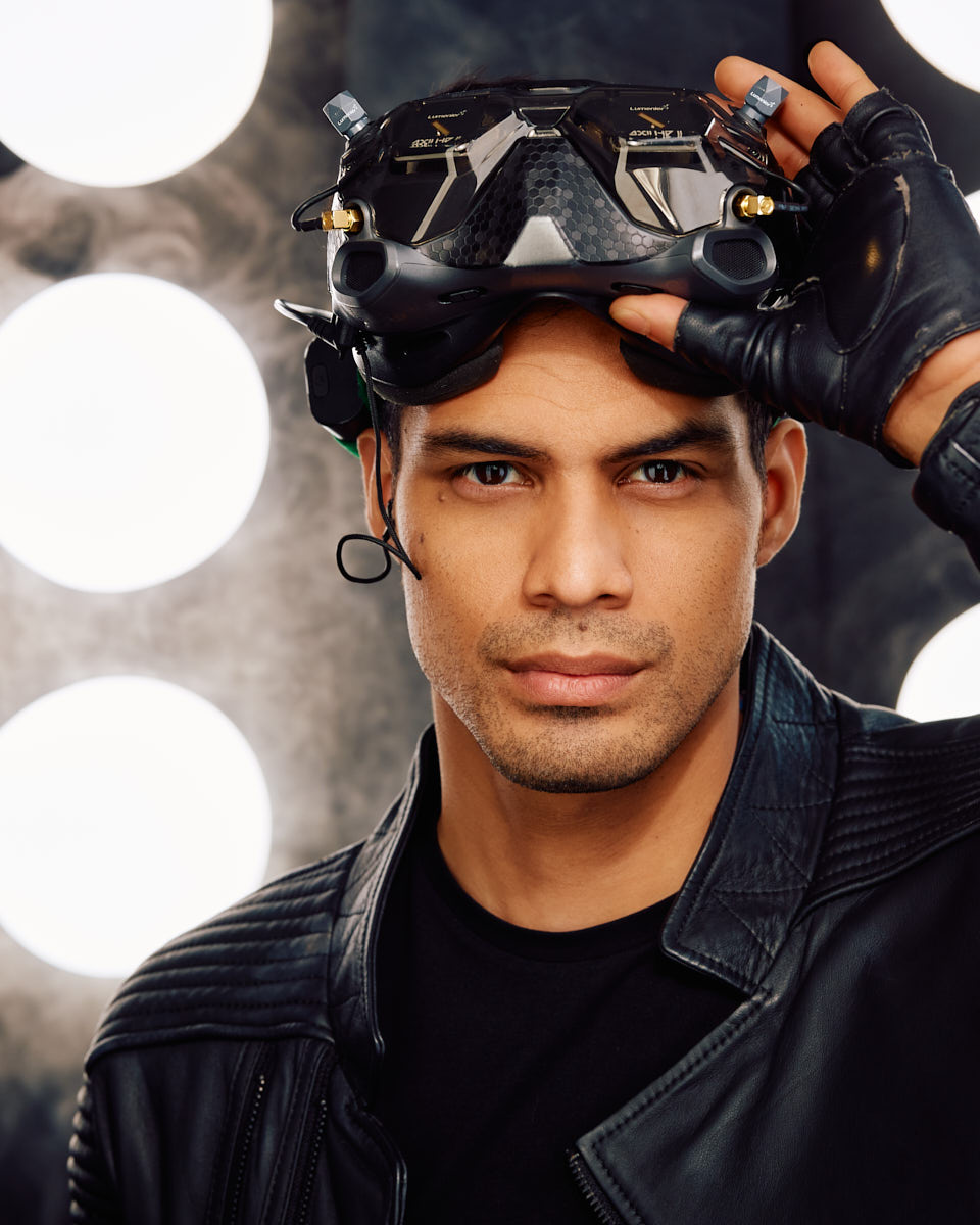 A male model in a leather jacket, holding a futuristic helmet, gazing intently at the camera against a background of circular lights.
