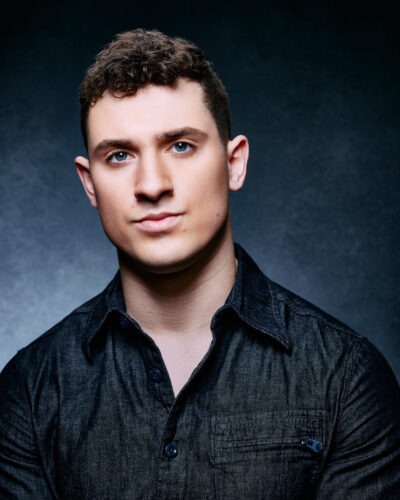 A close-up headshot of Tom with short curly hair and blue eyes, wearing a dark denim shirt, set against a dark background.