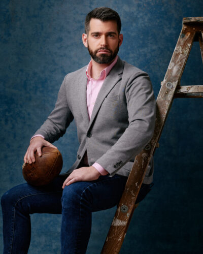 Jake is seated on a wooden ladder, holding a vintage football, and wearing a light grey blazer, pink shirt, and dark jeans. He has a focused expression and is looking directly at the camera, with a textured blue background adding depth to the image
