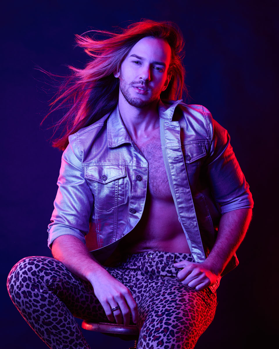 Glaydson Jr. is seated again, in a similar pose to the first image, but with a different angle that emphasizes his confident expression and the texture of his outfit against the vibrant background.