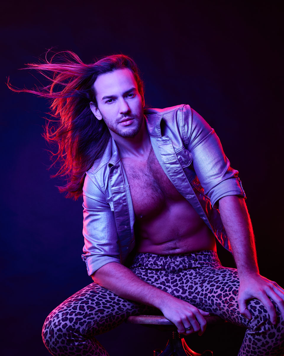 Male Model grooming tips in Chicago-Glaydson Jr. is posing with his long hair flowing and highlighted by vibrant, neon-like lighting. He is wearing a metallic jacket, partially open to reveal his chest, showcasing his well-groomed hair and glowing skin.