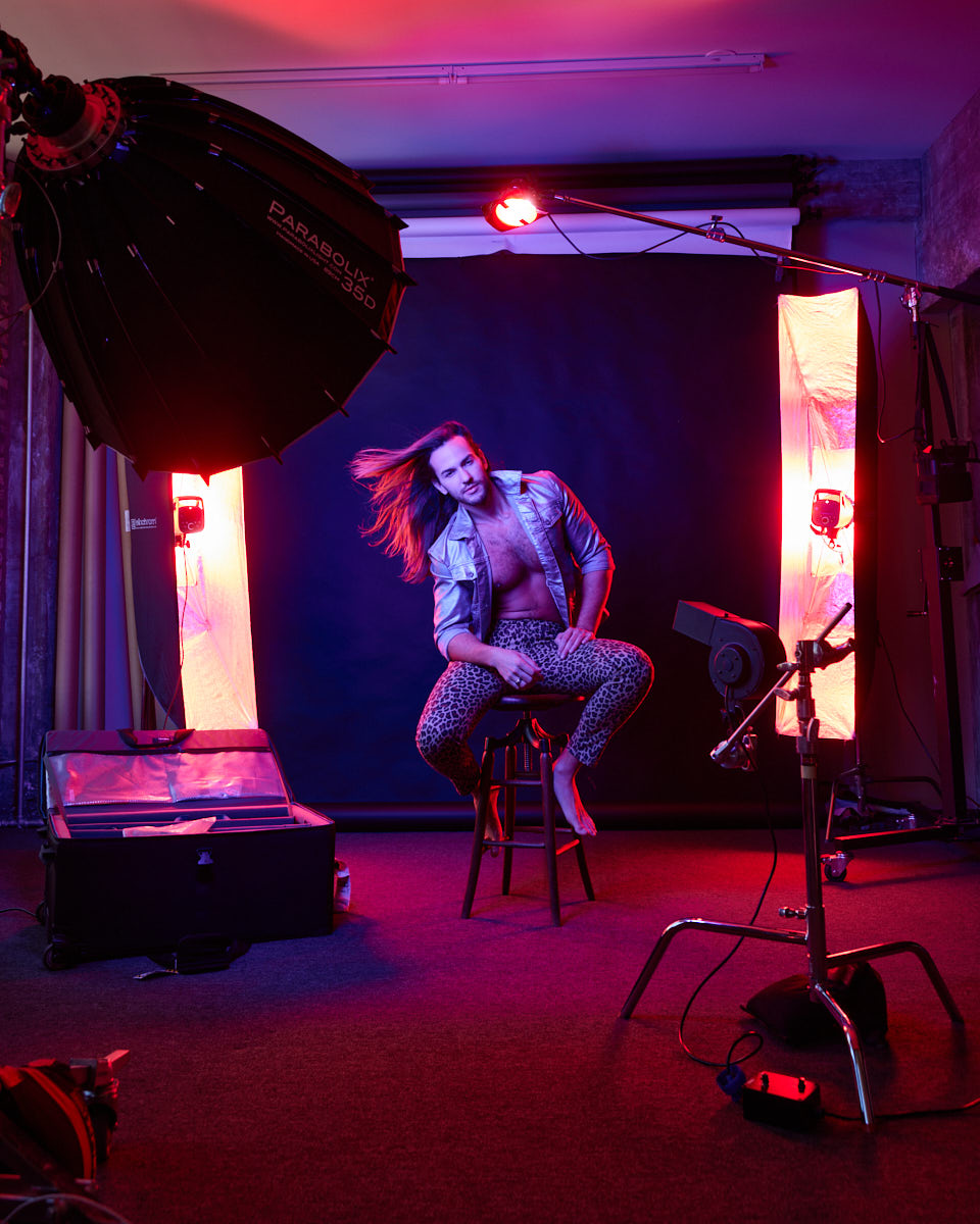 A behind-the-scenes shot of the photoshoot setup. Glaydson Jr. is seated on a stool with studio lights and equipment visible, giving a glimpse into the professional environment of the shoot.