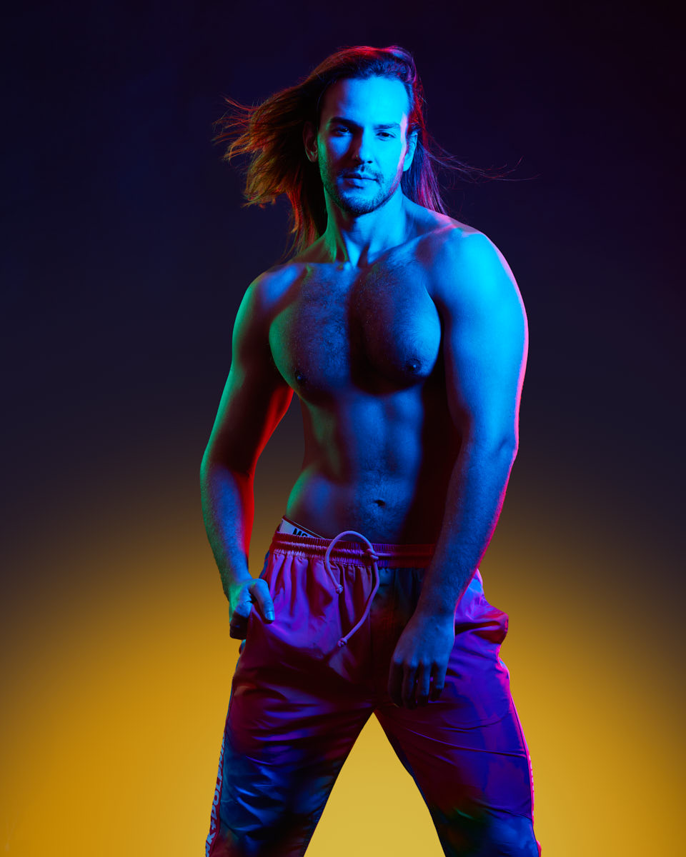 Male Model grooming tips in Chicago-Glaydson Jr. in a strong pose, shirtless, wearing colorful pants. The lighting casts a blue and red hue, creating a dramatic and eye-catching visual effect.