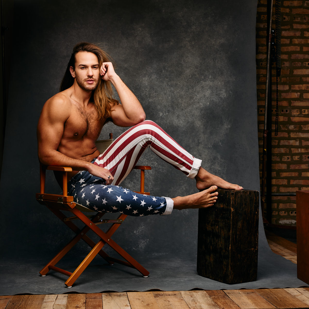 Glaydson Jr. seated in a director’s chair, wearing American flag-themed pants. His relaxed pose and the rustic studio setting create a contrast to the more vibrant images, showing his versatility as a model.