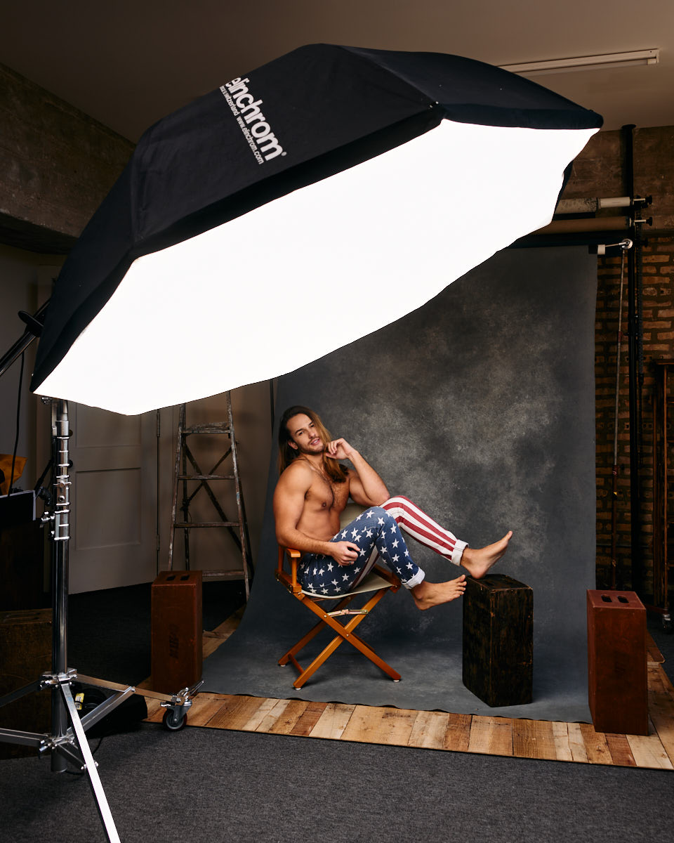 Male Model grooming tips in Chicago-In this behind-the-scenes image of the photoshoot, the male model Glaydson Jr. is seated in a director’s chair wearing American flag-themed pants, showcasing his relaxed yet confident pose. He is shirtless, and his hair is styled in a casual manner. The studio setup is visible, including a large softbox lighting equipment labeled “Elinchrom” prominently positioned to provide soft, even lighting for the shoot.