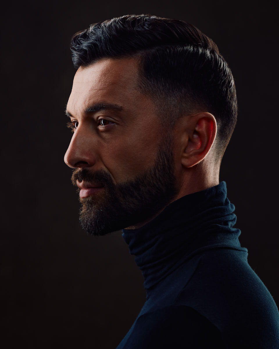 A color profile shot of Uros wearing a black turtleneck, against a dark background, showing his side profile.