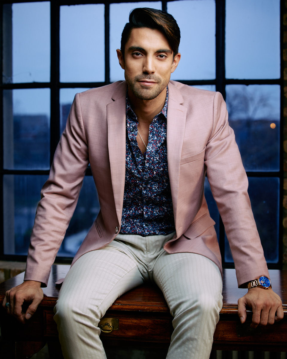 Male Model Headshots in Chicago-Daniel is sitting on a table, wearing a pink blazer, floral shirt, and light trousers. The backdrop is a window with an urban view, giving a modern and professional look.