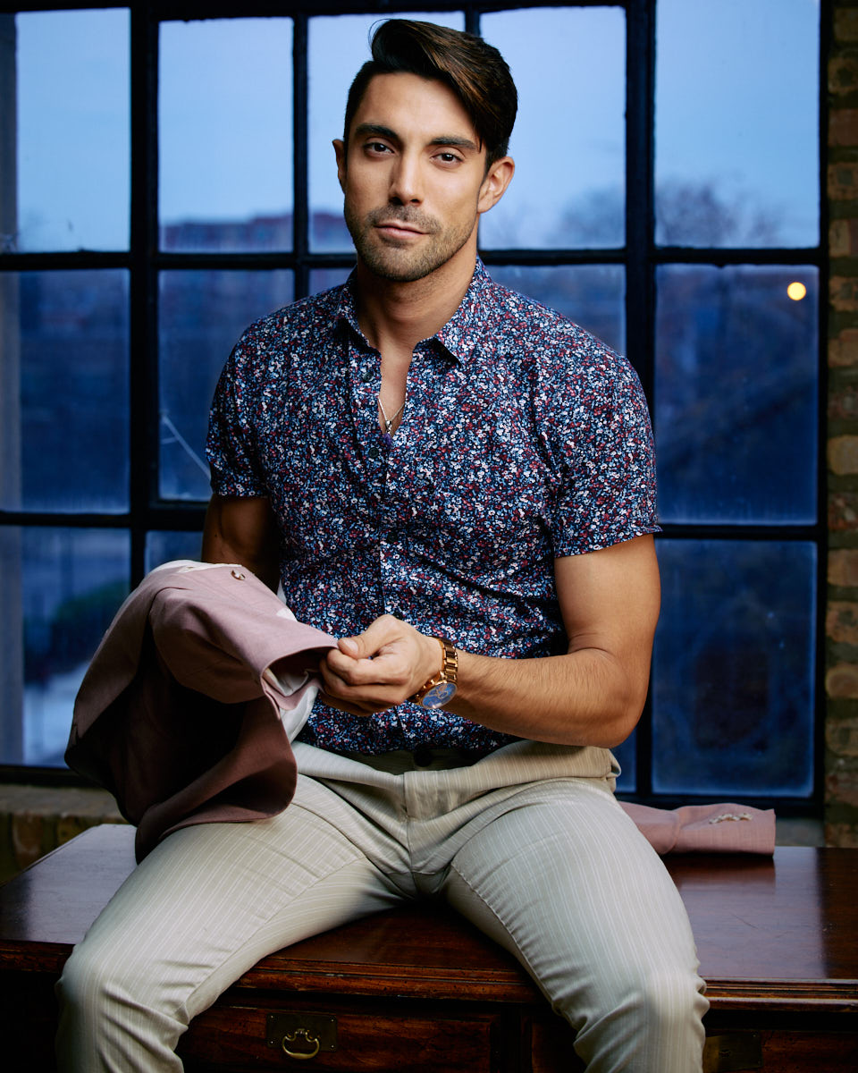 Male Model Headshots in Chicago-Daniel is sitting on a table in front of a window, wearing a floral shirt and light trousers, holding a pink blazer, with an urban backdrop.