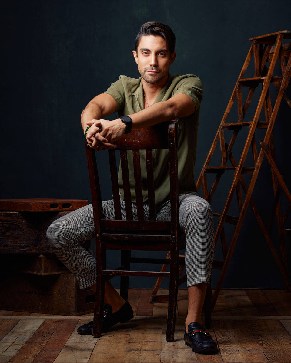 Daniel is seated, leaning on a wooden chair, wearing a green shirt and checkered pants. The setup includes wooden ladders and crates, creating a rustic studio environment.