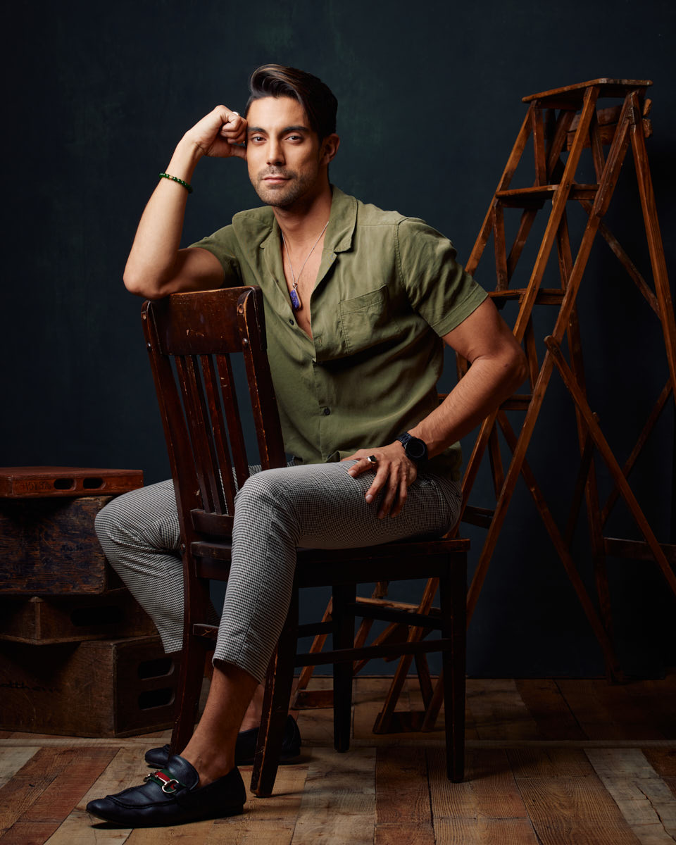 Daniel sitting in a green shirt, one arm resting on the chair back, looking confident and stylish in a rustic studio setting.