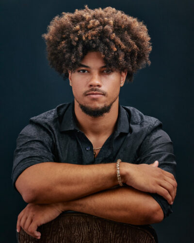 Josiah seated with his arms crossed, wearing a dark shirt, with a neutral studio background.
