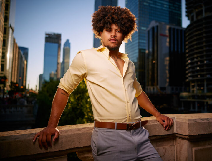 Josiah standing on a bridge in an urban setting, wearing a yellow shirt and light pants, with buildings in the background.
