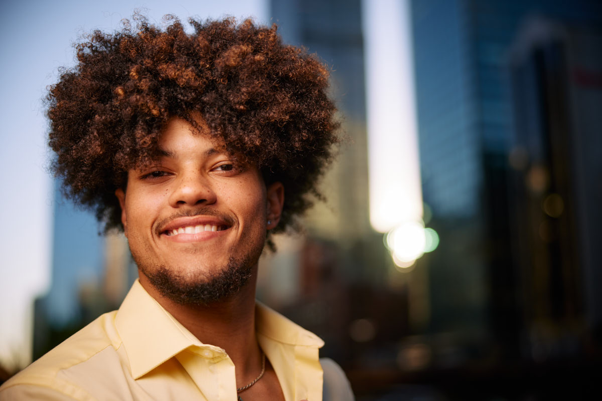 Josiah smiling broadly while standing outdoors in front of a cityscape, wearing a yellow shirt.
