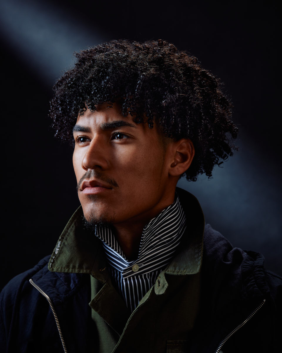 Kasan, a male model with curly hair, is posing against a dark background. A spotlight highlights his face. He is wearing a striped shirt with a high collar and a green jacket, giving a serious and focused expression